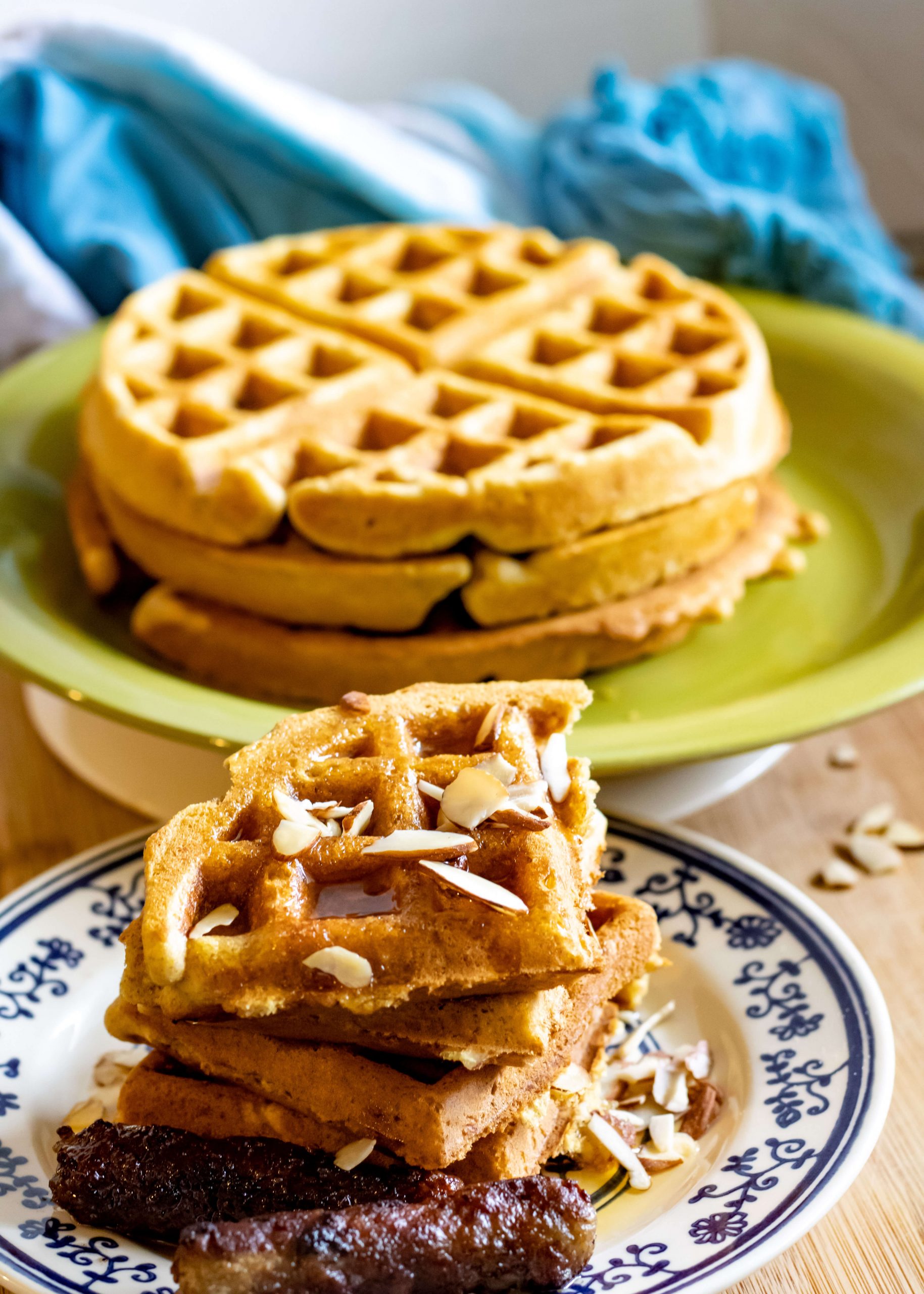 stack of waffles and a serving of belgian waffles topped with maple syrup and sliced almonds, served with 2 sausage links.