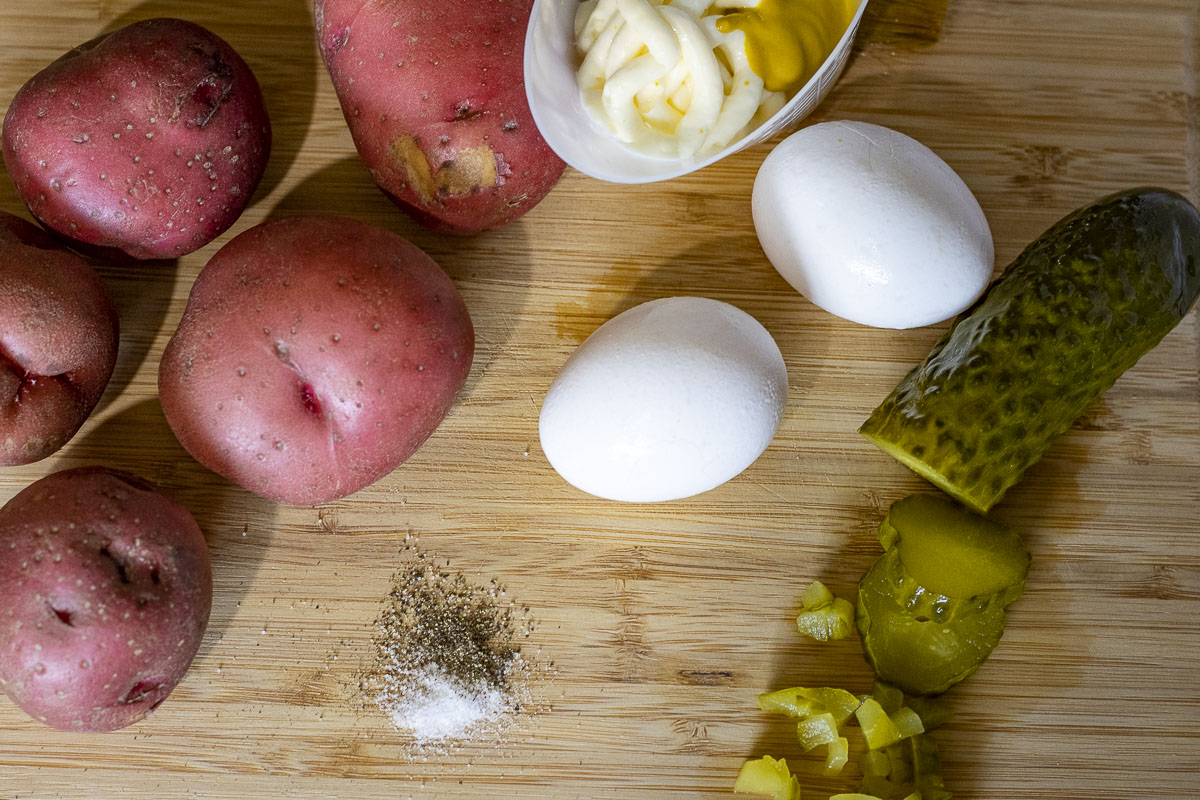 Ingredients for recipe on cutting board: whole red potatoes, mayo and mustard in dish, 2 boiled eggs, salt and pepper, and half a sliced pickle with some chopped.