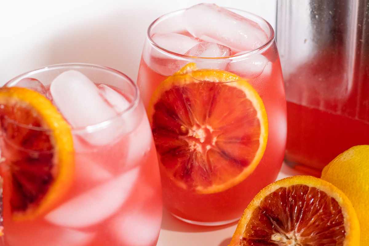 2 glasses of orange lemonade with ice next to a pitcher and extra fruit around.